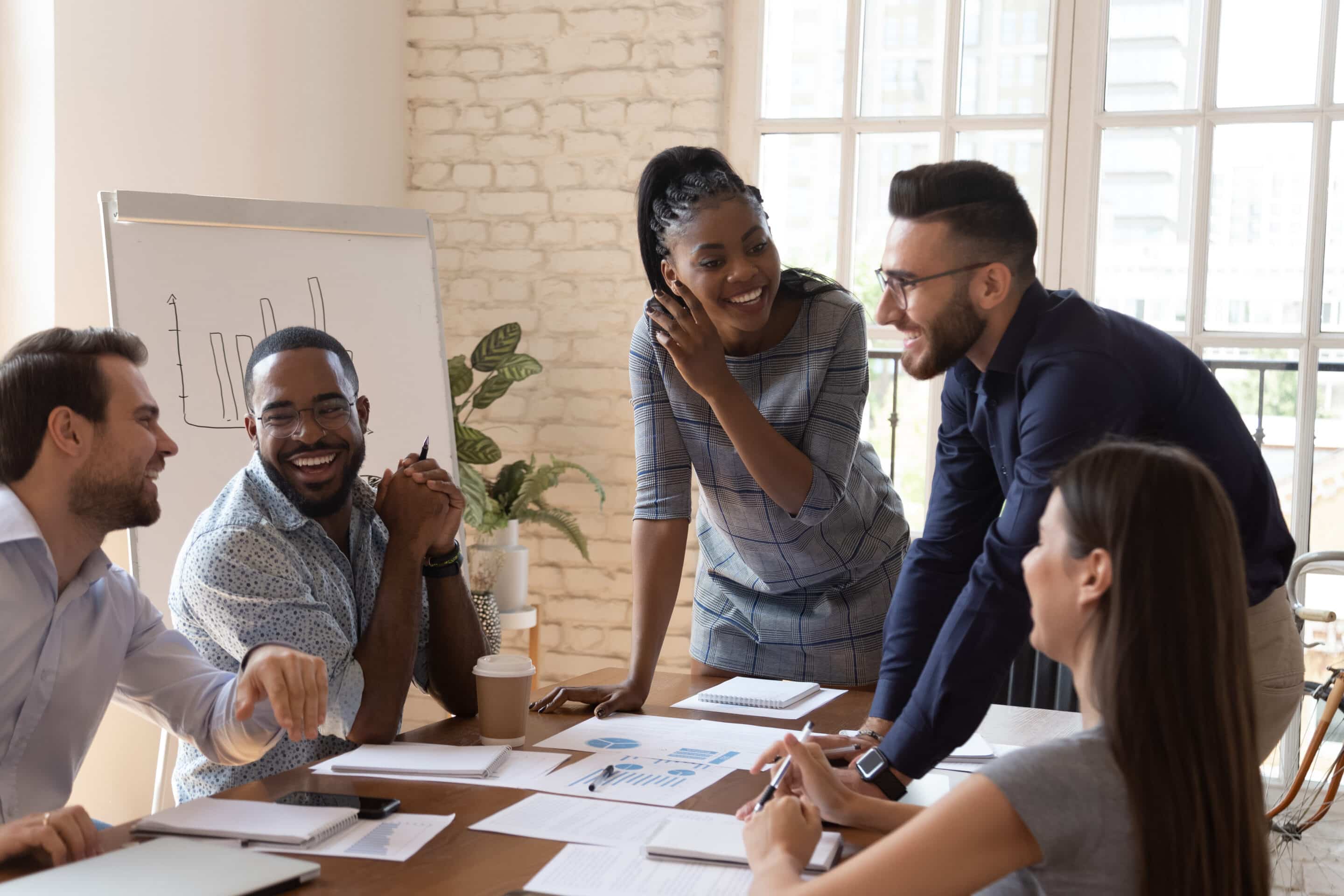 Happy friendly multiracial team laughing working together at corporate briefing Group of diverse coworkers collaborating around a conference table, reviewing charts and notes in a bright office.